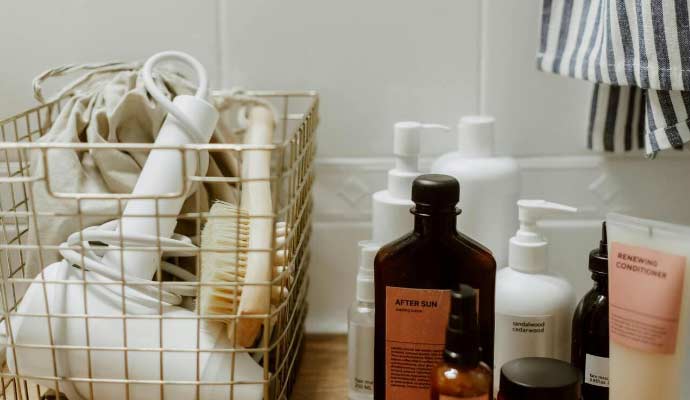 Metalna korpa sa fenom, četkom i platnenom torbom pored bočica kozmetike na kupatilskoj ploči