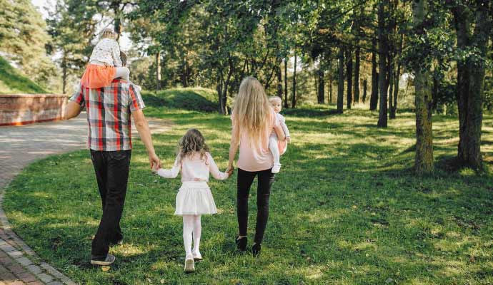 Porodica hoda kroz park, otac nosi dete na ramenima dok majka drži bebu
